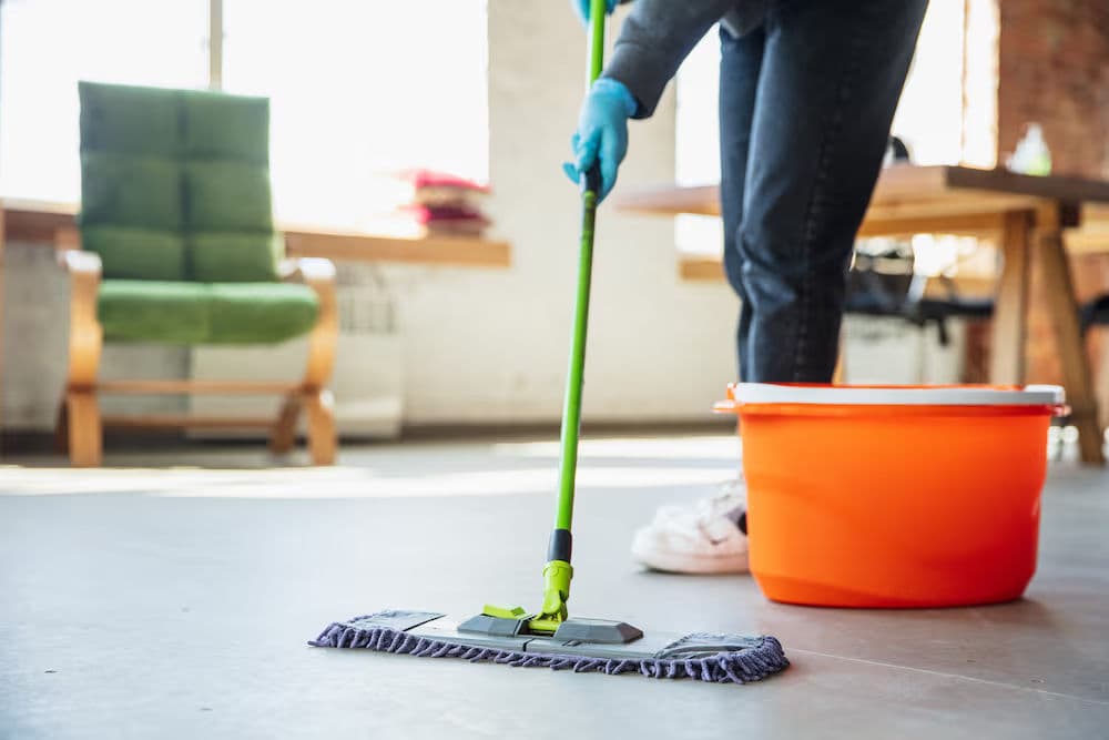 Person mopping a floor with a microfiber mop and orange bucket in a sunlit room.
